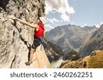 A woman is climbing a rock wall with a red tank top on. The mountain in the background is covered in trees