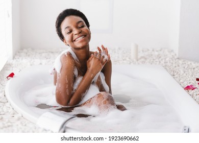 Woman with closed eyes of pleasure taking bath with foam and petals at home, spa, hygiene and relax. Smiling young cute african american woman enjoys warm water in interior in morning, copy space