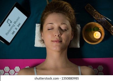 Woman with closed eyes Relaxing at home, lying on acupuncture mat. Candles and incense aroma sticks. Listening music from digital tablet