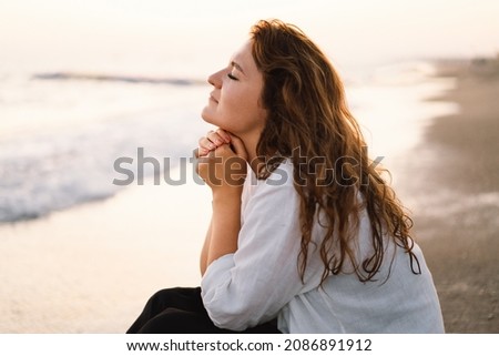 Woman closed her eyes, praying on a sea during beautiful sunset. Hands folded in prayer concept for faith, spirituality and religion. Peace, hope, dreams concept