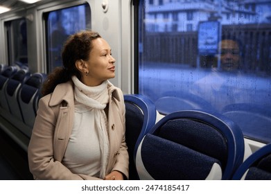 Woman in a coat and scarf sitting on a train, gazing thoughtfully out of the window on a rainy day, with her reflection visible. - Powered by Shutterstock