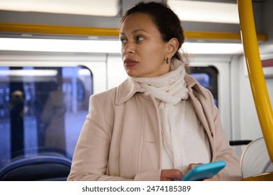 Woman in a coat and scarf using a smartphone while commuting on a public bus. She looks out the window in a thoughtful manner. - Powered by Shutterstock