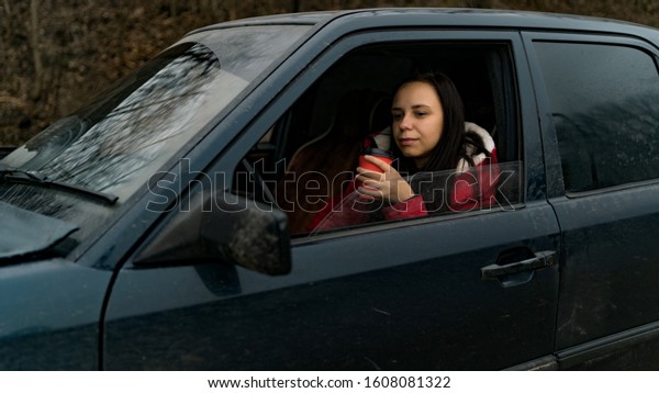 Woman\
with coffee in car looking out from window. Long haired woman with\
disposal cup at front seat of car looking at\
window