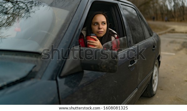 Woman\
with coffee in car looking out from window. Long haired woman with\
disposal cup at front seat of car looking at\
window