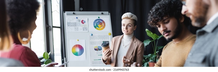 Woman With Coffee To Go Talking To Blurred Interracial Colleagues Near Flip Chart In Ad Agency, Banner