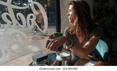 Woman In A Coffee Shop Looking Out The Window In A Nostalgic Scene, Watching The People Passing By, With A Cup Of Coffee In Her Hands, The Light Shining On Her Face. She Is Holding A Mobile Phone, 