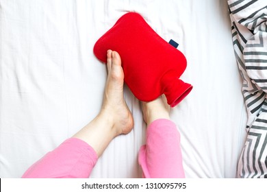 Woman With Cold Feet In Bed On A Red Hot Water Bottle. Warming Of Cold Feet Concept With Place For Text