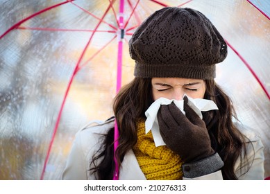 Woman With Cold Or Flu Coughing And Blowing Her Nose With A Tissue Under Autumn Rain. Brunette Female Sneezing And Wearing Warm Clothes.