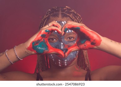 Woman with colorful face and hands paint and braids in her hair making a heart gesture in front of her eyes with her hands. Isolated on a background with red smoke. - Powered by Shutterstock