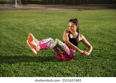 A woman in colorful geometric leggings and a black sports top performs a core rotational exercise on a grassy field, showcasing strength and focus during an outdoor fitness session - Powered by Shutterstock