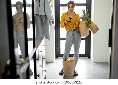 Woman Comes Home With Bags Full Of Fresh Groceries, Standing With Phone At The Hallway. Concept Of Buying Food Online And Delivering It Home