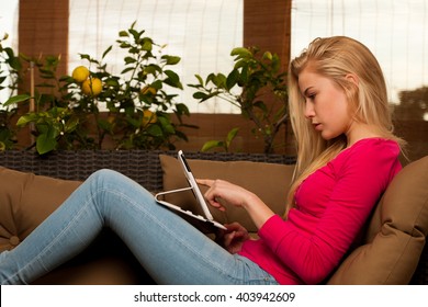 Woman comfortable sitting on sofa surrounded with lemon tree and other green plants and using tablet computer.