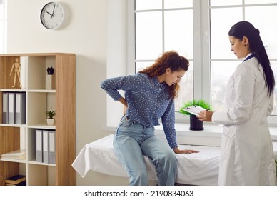 Woman Complains To Doctor About Back Or Kidney Pain During Medical Examination In Hospital Room. Female Doctor Records Complaints Of Patient Who Is Holding Her Back While Sitting On Medical Couch.