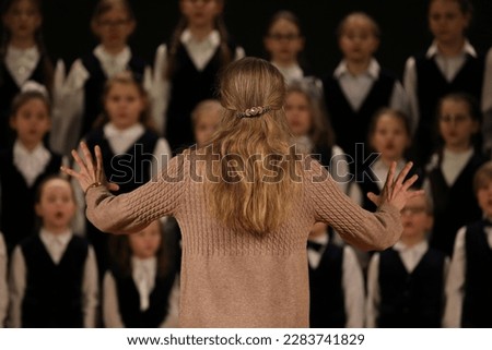 A woman conducts children singing chorus in action active energetic leader back view hands up
