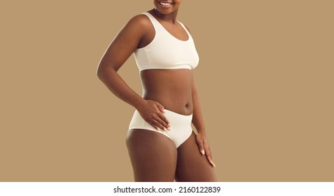 Woman is confident in her beauty and enjoys her size and type of figure standing on beige background. Cropped image of black woman in white cotton underwear who has no complexes about her appearance.