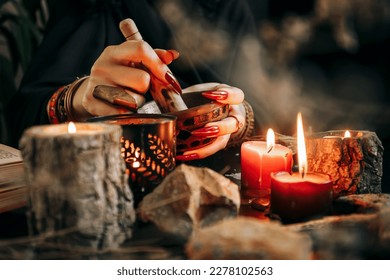 A woman conjures and grinds dried plants in a wooden mortar among lighted candles against a dark background - Powered by Shutterstock