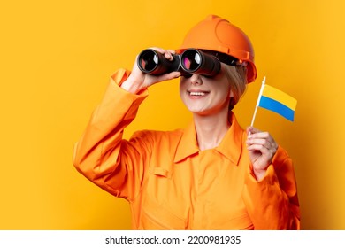 Woman Construction Worker In Helmet And Orange Overalls With Binoculars And Ukrainian Flag On Yellow Background