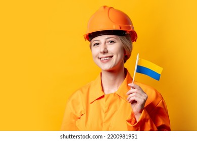 Woman Construction Worker In Helmet And Orange Overalls Holding Ukrainian Flag Against A Yellow Background 