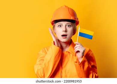 Woman Construction Worker In Helmet And Orange Overalls Holding Ukrainian Flag Against A Yellow Background 