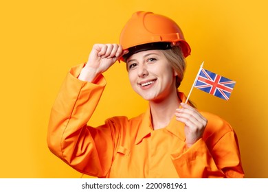 Woman Construction Worker In Helmet And Orange Overalls Holding GB Flag Against A Yellow Background