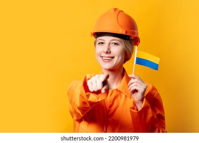 Woman Construction Worker In Helmet And Orange Overalls Holding Ukrainian Flag Against A Yellow Background 