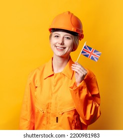 Woman Construction Worker In Helmet And Orange Overalls Holding GB Flag Against A Yellow Background