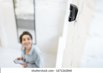 Woman controlling alarm system with a smart phone wireless, standing in the room with motion sensor mounted on the wall
