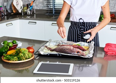 Woman Cooking Trout Fish Using The Recipe A Tablet Computer To Cook In Her Kitchen.