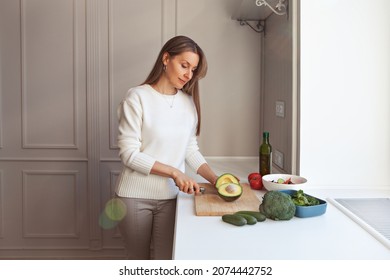 Woman cooking vegetarian salad with fresh vegetables. Model cutting with knife avocados, broccoli, cale salad, cucumbers, tomatoes in white kitchen