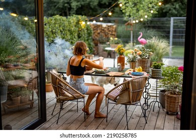 Woman Cooks Food On Disposable Grill While Sitting Relaxed By The Table On Cozy Terrace During The Evening At Beautiful Backyard