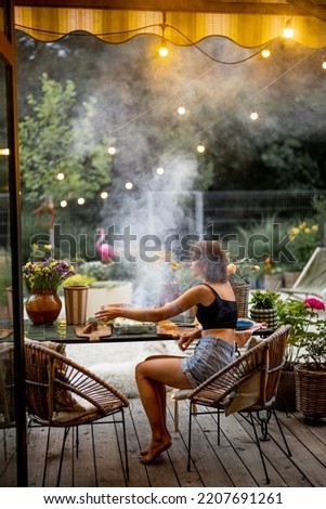 Woman cooks food on disposable grill while sitting relaxed by the table on cozy terrace during the evening at beautiful backyard