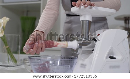 Woman cooks sausages in traditional way using sausage filler of electric mincer machine in kitchen. Production of meat delicacies at home. Preparation of raw beef sausages with electric grinder.