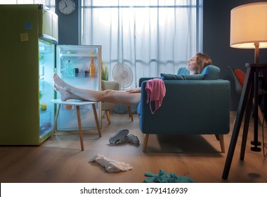Woman cooling herself in front of the open fridge at home during the summer, she is sitting on the sofa with feet up - Shutterstock ID 1791416939