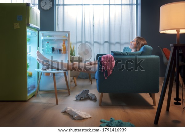 Woman\
cooling herself in front of the open fridge at home during the\
summer, she is sitting on the sofa with feet\
up