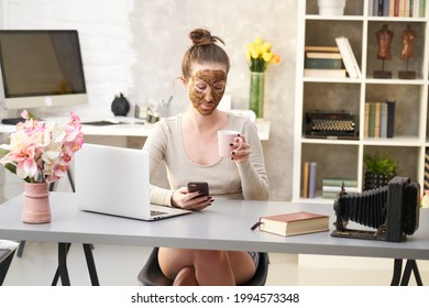 Woman Cosmetic Facial Skin Care. Woman In Skin Rubber Peeling Face Beauty Mask Sitting At Desk Using Phone And Laptop In Home Office.