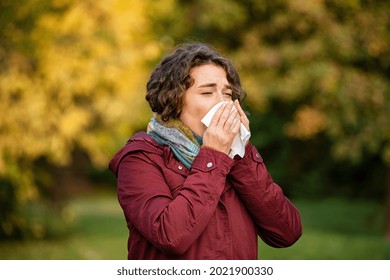 Woman coughing and blowing her nose in park during autumn season. Sick young woman with nose wiper caught cold. Girl blowing in a tissue in a cold autumn day.