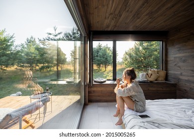 Frau in einem Landhaus oder Hotel mit Panoramafenstern in Kiefernwald sitzend auf dem Bett und genießt eine schöne Aussicht mit leichter Sonne. Guten Morgen und Erholung am Naturkonzept