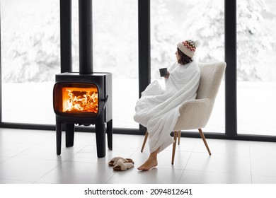 Woman Covered With Blanket Enjoys Winter Time At Home With Burning Fireplace, Looking Outside The Window On Snowy Landscape. Concept Of Winter Mood. Idea Of Winter Vacation In The Mountains