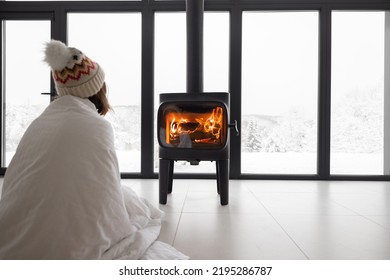 Woman covered with blanket enjoys winter time at home with burning fireplace, looking outside the window on snowy landscape. Concept of winter mood. Idea of winter vacation in the mountains