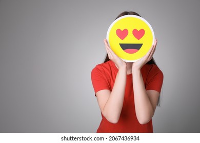 Woman covering face with heart eyes emoji on grey background, space for text