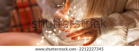 woman in a cozy comfy knitted wool white sweater holding in hands and untangle glowing lights garland at home on the couch or bed. new year and christmas eve decorating and celebration. banner