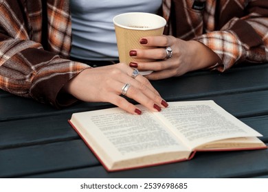 Woman in a cozy flannel shirt reads a book and enjoys coffee outdoors at a dark wooden table, surrounded by autumn tranquility - Powered by Shutterstock