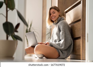 Woman in cozy home clothes relaxing at home, using laptop. Beautiful girl in knitted warm grey sweater and knitted socks sitting on the floor and working on laptop and smiling. Comfy lifestyle.