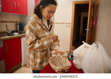 A woman in a cozy plaid jacket organizes groceries in a kitchen while a child plays in the background. The warm and inviting atmosphere highlights a daily domestic scene. - Powered by Shutterstock