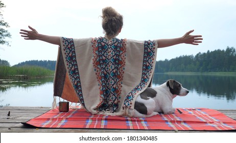 Woman in cozy poncho wide spreading her hands appreciating the nature and her time with white and brown dog on the dock of the lake at sunset. Self care concept.  - Shutterstock ID 1801340485