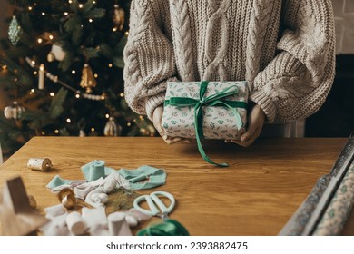 Woman in cozy sweater holding stylish christmas gift in festive wrapping paper with bow, ribbon, vintage ornaments on wooden table. Atmospheric winter holidays, wrapping christmas present - Powered by Shutterstock