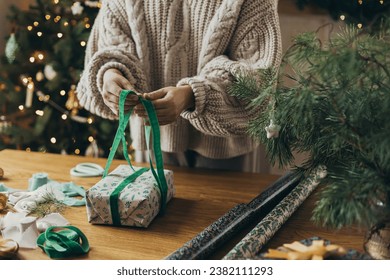 Woman in cozy sweater wrapping stylish christmas gift in festive wrapping paper with ribbons, vintage ornaments, bows on wooden table. Atmospheric winter holidays, wrapping christmas present - Powered by Shutterstock