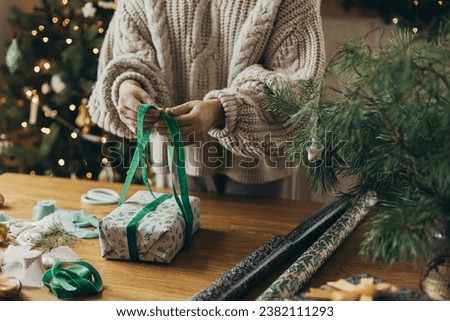 Woman in cozy sweater wrapping stylish christmas gift in festive wrapping paper with ribbons, vintage ornaments, bows on wooden table. Atmospheric winter holidays, wrapping christmas present