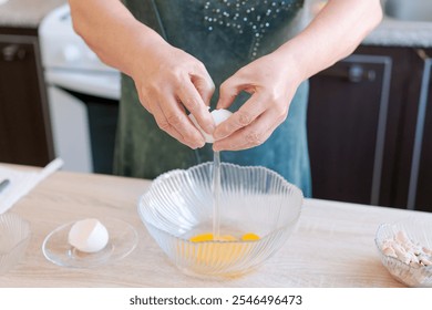 Woman is cracking an egg into a bowl. The bowl is on a wooden table. The woman is wearing a green apron - Powered by Shutterstock