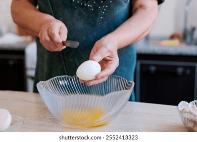 Woman is cracking an egg into a bowl. She is wearing a green shirt. The bowl is on a wooden table - Powered by Shutterstock